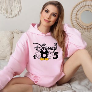Op de afbeelding: Roze hoodie met een zwarte en gele Mickey Mouse-afbeelding en de tekst "Disney 5".