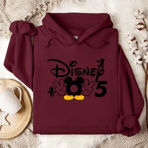 Op de afbeelding: Bordeauxkleurige hoodie met een zwarte en gele Mickey Mouse-afbeelding en de tekst "Disney 5".