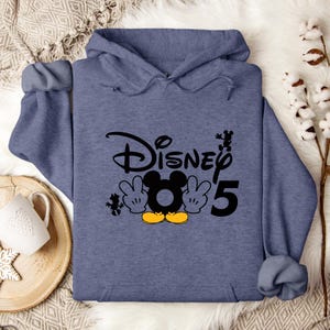 Op de afbeelding: Een blauw hoodie met de tekst "Disney" en het nummer "5" in het zwart. Een cartoon afbeelding van Mickey Mouse met gele handschoenen en schoenen bevindt zich in het midden van de tekst.