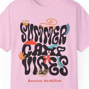 May include: Pink t-shirt featuring a black graphic that reads "Summer Camp Vibes" in a retro font. The graphic includes colourful illustrations of flowers, mountains, and a sun. The text "Kerrian McMillan" is printed below the graphic.