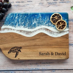 May include: Wooden serving board with a handle, featuring a beach-themed design with blue resin waves and a sand-colored base. Includes a turtle engraving and the names "Sarah & David".