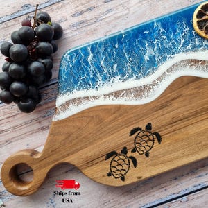 May include: Wooden serving board with a beach-themed design. Features a blue and white resin wave pattern, a sandy-colored wood base, and two turtle silhouettes. Includes a handle with a circular opening.