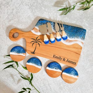 May include: A wooden serving board with a handle, featuring a blue resin ocean wave design, a black palm tree silhouette, and the text Emily & James. It includes four matching round wooden coasters with blue resin waves and four metal cheese knives.