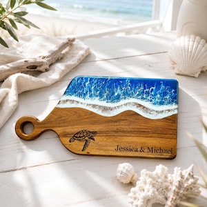 May include: A wooden serving board with a handle featuring a resin ocean wave design in shades of blue and white. The board includes a black engraved sea turtle illustration and the text "Jessica & Michael" on the natural wood surface.