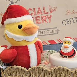 May include: A plush yellow duck wearing a Santa Claus outfit, including a red hat with white trim and a red coat with white trim. A smaller Santa duck figurine sits on a white pedestal. The background features a Christmas-themed design.