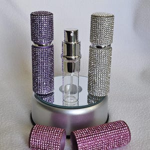 Puede incluir: Una colecci&oacute;n de atomizadores de perfume con adornos de pedrer&iacute;a. Los atomizadores son de color p&uacute;rpura, plateado y rosa. Un atomizador est&aacute; abierto, revelando el frasco de vidrio. Los atomizadores se exhiben sobre una superficie espejada.