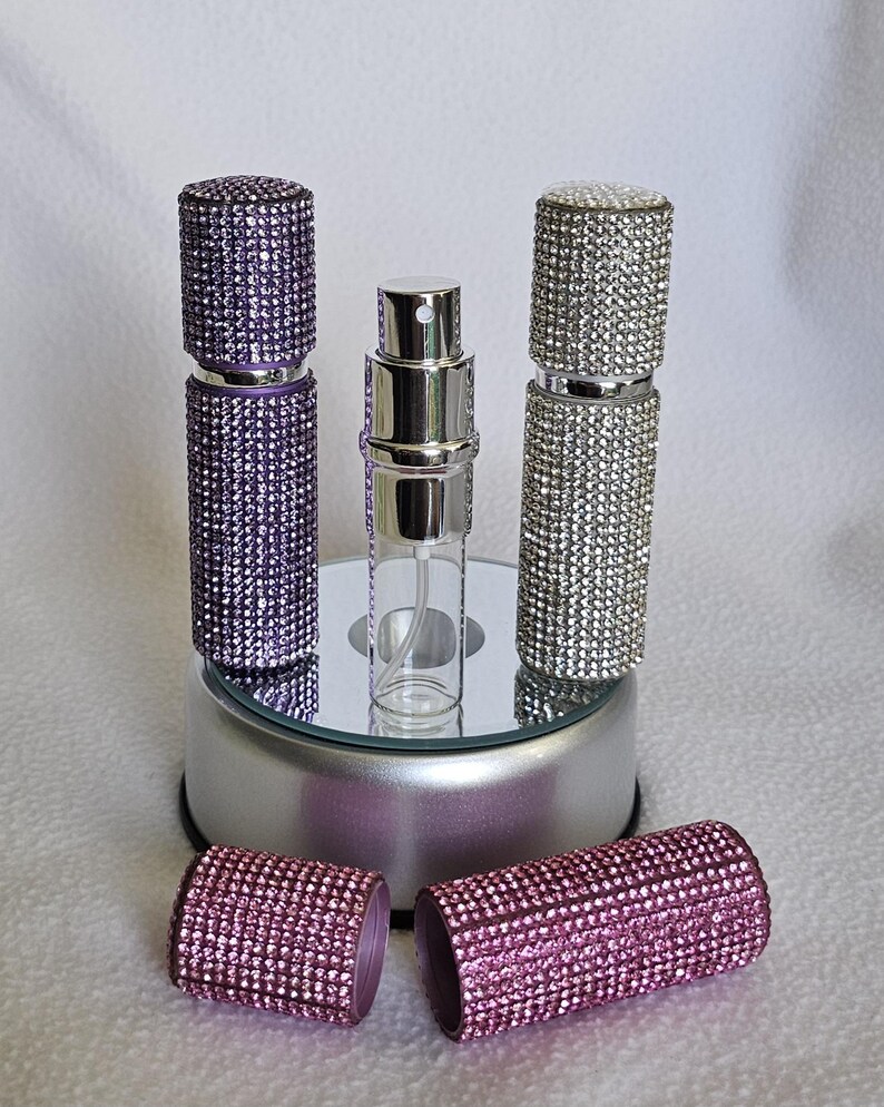 Puede incluir: Una colecci&oacute;n de atomizadores de perfume con adornos de pedrer&iacute;a. Los atomizadores son de color p&uacute;rpura, plateado y rosa. Un atomizador est&aacute; abierto, revelando el frasco de vidrio. Los atomizadores se exhiben sobre una superficie espejada.