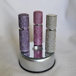 Puede incluir: Tres atomizadores de perfume enjoyados en morado, rosa y plateado. Cada atomizador est&aacute; cubierto de peque&ntilde;os y brillantes pedrer&iacute;a. Los atomizadores se exhiben sobre una superficie espejada encima de una plataforma plateada.