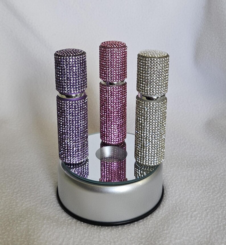 Puede incluir: Tres atomizadores de perfume enjoyados en morado, rosa y plateado. Cada atomizador est&aacute; cubierto de peque&ntilde;os y brillantes pedrer&iacute;a. Los atomizadores se exhiben sobre una superficie espejada encima de una plataforma plateada.
