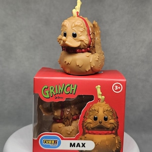May include: A brown Max figure from the Grinch movie, atop a red box. The figure has a red collar and a yellow antler. The box reads "Grinch" and "Max" with a "3+" age rating.