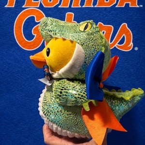 Könnte beinhalten: Eine Plüsch-Ente in einem grünen Alligator-Kostüm mit einer blauen und orangefarbenen Schleife. Die Ente ist gelb mit einem orangefarbenen Schnabel, vor einem blauen Hintergrund mit den Worten "FLORIDA Gators" in Orange.