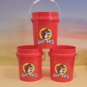 May include: Three red plastic buckets stacked. Each bucket has a white handle and a circular logo with a cartoon beaver and the text "BUC-EE'S". The buckets are cylindrical with a slightly flared top.