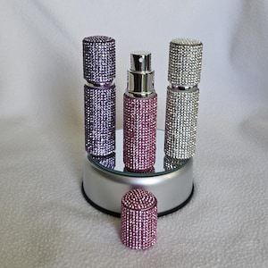 Puede incluir: Una colecci&oacute;n de atomizadores de perfume enjoyados en morado, rosa y plateado. Los atomizadores son cil&iacute;ndricos con una boquilla rociadora y est&aacute;n cubiertos de peque&ntilde;os pedrer&iacute;a brillantes. Un atomizador rosa tiene la tapa quitada y se coloca delante de los dem&aacute;s.