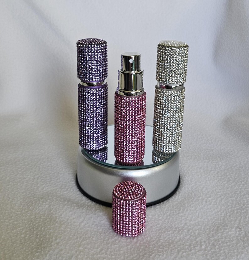 Puede incluir: Una colecci&oacute;n de atomizadores de perfume enjoyados en morado, rosa y plateado. Los atomizadores son cil&iacute;ndricos con una boquilla rociadora y est&aacute;n cubiertos de peque&ntilde;os pedrer&iacute;a brillantes. Un atomizador rosa tiene la tapa quitada y se coloca delante de los dem&aacute;s.