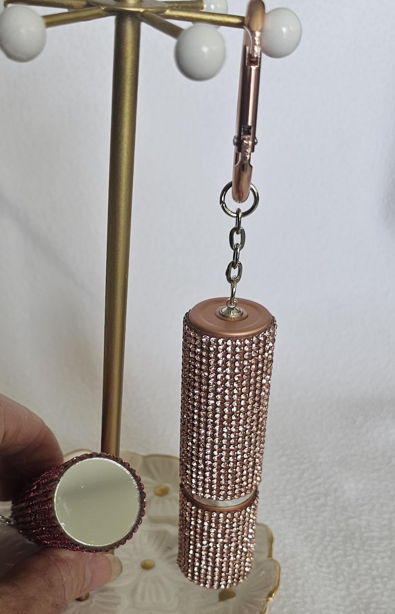 Puede incluir: Un estuche de l&aacute;piz labial de oro rosa incrustado con peque&ntilde;os pedrer&iacute;a brillantes. El estuche de l&aacute;piz labial est&aacute; sujeto a una cadena y un clip de oro rosa. Tambi&eacute;n se ve un peque&ntilde;o espejo redondo de oro rosa.