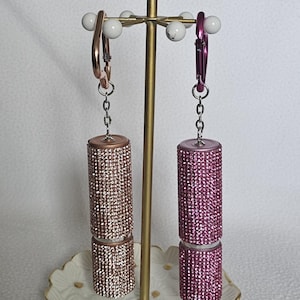 Puede incluir: Un expositor de joyas decorativo con dos recipientes con forma de l&aacute;piz labial adornados con pedrer&iacute;a. Uno es oro rosa, el otro rosa. El expositor tiene un tallo dorado y una parte superior floral blanca, sobre un plato floral blanco y dorado.