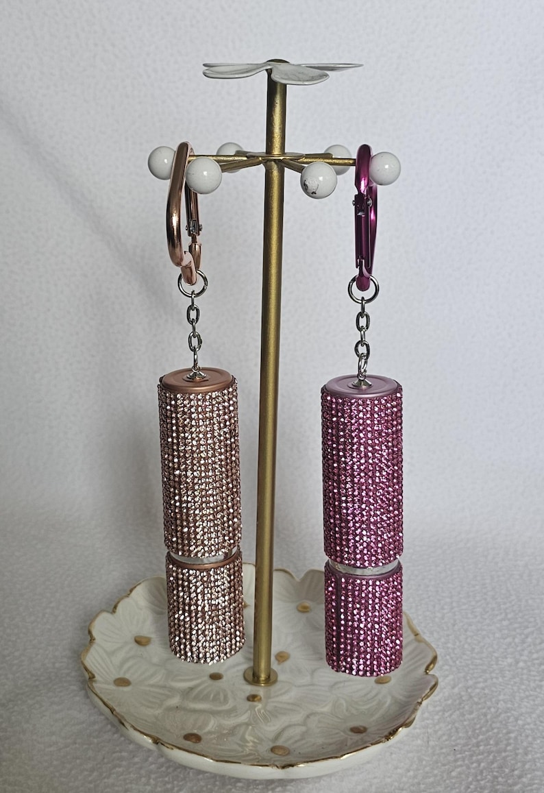 Puede incluir: Un expositor de joyas decorativo con dos recipientes con forma de l&aacute;piz labial adornados con pedrer&iacute;a. Uno es oro rosa, el otro rosa. El expositor tiene un tallo dorado y una parte superior floral blanca, sobre un plato floral blanco y dorado.