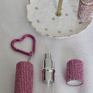 Puede incluir: Un conjunto de accesorios de belleza, que incluye un frasco de perfume rosa cubierto de pedrer&iacute;a, un labial a juego y un llavero en forma de coraz&oacute;n. Un plato blanco con estampado floral y detalles dorados sostiene el labial. Los art&iacute;culos est&aacute;n dispuestos sobre una superficie blanca.