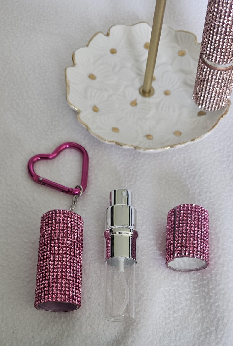 Puede incluir: Un conjunto de accesorios de belleza, que incluye un frasco de perfume rosa cubierto de pedrer&iacute;a, un labial a juego y un llavero en forma de coraz&oacute;n. Un plato blanco con estampado floral y detalles dorados sostiene el labial. Los art&iacute;culos est&aacute;n dispuestos sobre una superficie blanca.