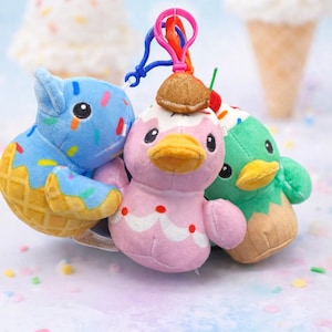 May include: Three plush duck keychains in the image. One is blue with a waffle cone base, another is pink with a cake design, and the third is green with a waffle cone base. Each has a colorful clip for attachment.