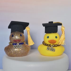Könnte beinhalten: Zwei Gummienten, dekoriert für eine Abschlussfeier. Eine ist goldglitzernd, die andere gelb. Beide tragen schwarze Mortarboards mit goldenen Quasten und Perlenketten mit einem "2026"-Anhänger. Blaue Augen und orangefarbene Schnäbel.
