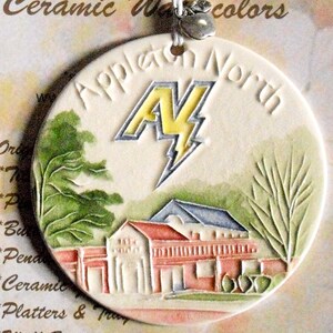 Könnte beinhalten: Ein rundes Keramikornament mit einem Reliefdesign eines Gebäudes mit rotem Dach und grünen Bäumen. Der Text "Appleton North" ist über einem gelb-silbernen Blitz-Logo eingeprägt. Ein kleiner silberner Apfelanhänger hängt an der Oberseite.