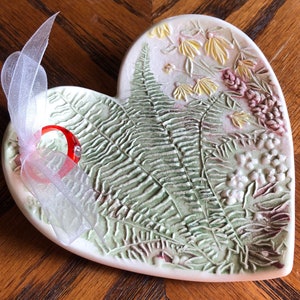May include: A heart-shaped ceramic dish featuring a raised, textured relief of green fern fronds and small yellow and pink flowers. A translucent red ring is attached to the left side of the dish with a sheer white ribbon tied in a bow.