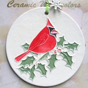 May include: A round, off-white ceramic ornament featuring a red cardinal bird perched on green holly leaves. The ornament has a green string for hanging and a silver snowflake charm. The text "Ceramic Watercolors" is at the top.