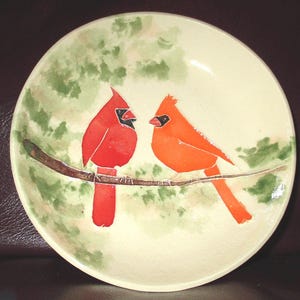 May include: A round ceramic plate with a cream-coloured base featuring a hand-painted illustration of two cardinal birds perched on a brown branch. The bird on the left is bright red, while the bird on the right is orange, set against green foliage.