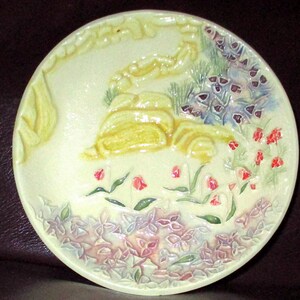 May include: A round, pale yellow ceramic plate featuring a raised, embossed design of a winding path, green foliage, and clusters of small purple, pink, and red flowers. The relief texture creates a three-dimensional garden scene on the surface.