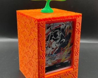 Op de afbeelding: Oranje en groene 3D-geprinte kaarthouder met een decoratief ontwerp. De houder heeft een transparant venster om een ​​verzamelkaart te tonen. De getoonde kaart is een "Sabo"-kaart uit het One Piece Trading Card Game. De kaart heeft een krachtniveau van 5000.