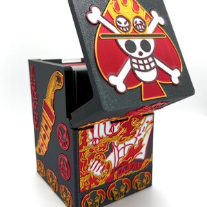 May include: Black box with a hinged lid, featuring a red and gold Ace of Spades design with a skull and crossbones. The box has additional gold and red accents, including a sword and skull motifs, and a fiery design on the inside.