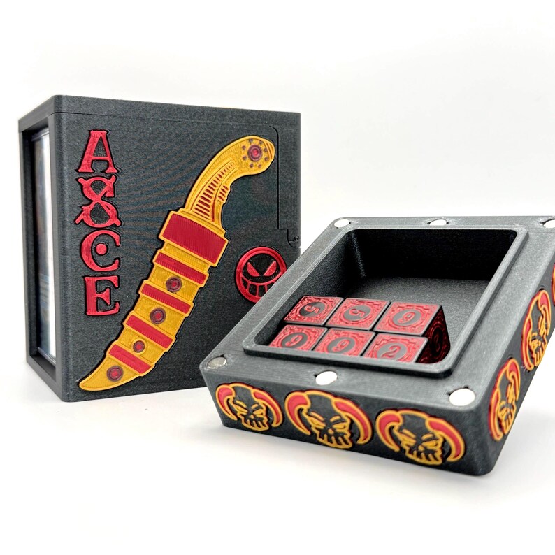 May include: A black dice box and tray set with red and yellow accents. The box features the word "AXCE" and a stylized knife design. The tray holds six red dice with white numbers and is decorated with skull motifs.