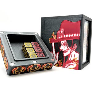 May include: A black and gray dice box with a red and gold design, open to reveal dice. A matching black box with a red and white character design is next to it. The dice are red, gold, and yellow.
