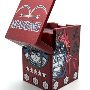 May include: A red card storage box with a hinged lid, featuring the word "MARINE" and a stylized emblem. The box is decorated with character illustrations, stars, and floral designs. The box is open, revealing the interior.