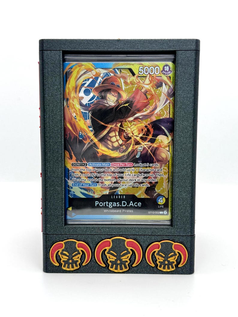 May include: A black card holder with a clear window displaying a trading card. The card features a character with a hat and flames, with text that reads "Portgas.D.Ace". The holder has three skull and flame designs.