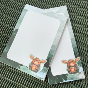 May include: Two white notepads with a watercolor forest design border. Each notepad features a cartoon moose holding a coffee cup. The notepads are stacked on a green woven surface.
