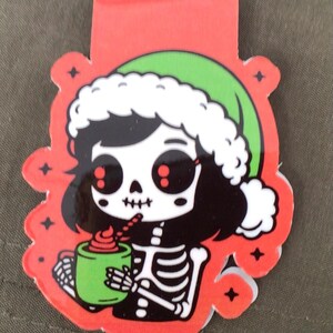 May include: A red and green Christmas-themed bookmark featuring a skeleton character wearing a Santa hat and holding a green mug with a drink. The bookmark has a red top and a decorative border with star accents.