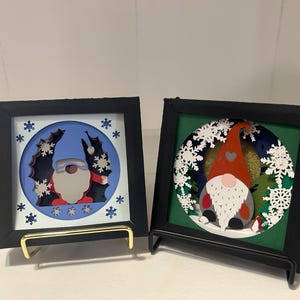 May include: Two framed Christmas-themed art pieces. Each features a gnome design within a circular frame, surrounded by snowflakes. One has a blue background, the other green. Both are in black frames with gold stands.