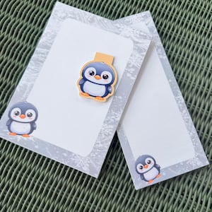 Puede incluir: Dos tarjetas rectangulares blancas con borde gris de copo de nieve y detalles de pingüino de dibujos animados. Una tarjeta tiene un marcapáginas con forma de pingüino adjunto. La otra tarjeta tiene una pegatina de pingüino en la esquina. El fondo es una textura tejida verde.