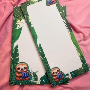 May include: Two rectangular notepads with a white interior and a green border featuring tropical leaves. Each notepad has a cartoon sloth reading a book. The background is a pink fabric.
