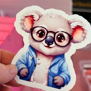 May include: A sticker featuring a cartoon koala wearing black glasses and a blue jacket. The koala has a white face, gray fur, and large red eyes. The sticker is cut around the koala's outline.