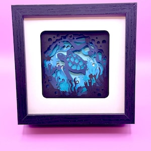 May include: A framed, layered paper art piece depicting an underwater scene. The artwork features a sea turtle, coral, and other aquatic elements in shades of blue and teal. The frame is dark wood, and the background is a soft pink.