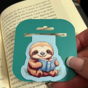 May include: A teal bookmark featuring a cartoon sloth reading a blue book. The sloth is brown and tan with large eyes and a smile. The bookmark has a rectangular shape with a rounded top and a hole for hanging.