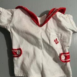 May include: White sailor suit with red trim. The top has a red collar and a small pocket with an embroidered anchor. The shorts have red accents and buttons. The outfit is made of a textured fabric.