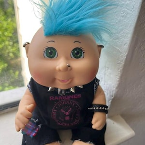 May include: A doll with bright blue hair styled in a mohawk, green eyes, and a nose ring. It wears a black sleeveless shirt featuring the text RAMONES and JOHNNY DEE DEE TOMMY, a silver spiked collar, a black studded wristband, and black pants.