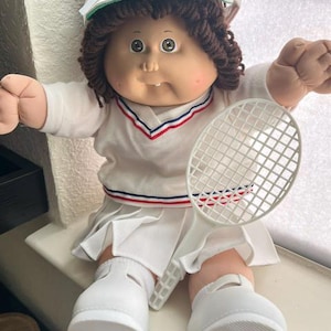 May include: A doll with brown yarn hair wearing a white visor, a white V-neck sweater with red and blue trim, and a white pleated skirt. The doll has white shoes and holds a white plastic tennis racket. The doll features a single tooth.