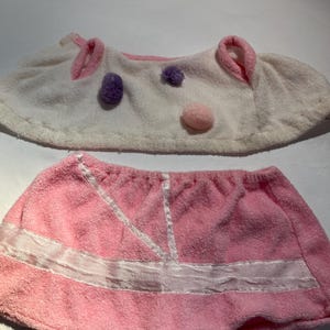 May include: Two terry cloth hair wraps and a skirt. The top wrap is white with pink trim and pom-poms. The bottom skirt is pink with white ribbon accents. The items appear to be for use after bathing or showering.