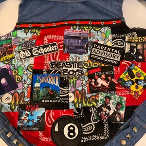 May include: A denim dog vest with a patchwork design featuring music album covers, graffiti art, and the text "Parental Advisory Explicit Content." The vest has metal stud accents and a leather tag with a skull and crossbones.