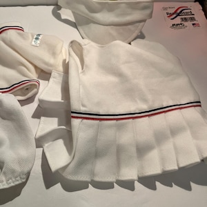 May include: A white tennis outfit set, including a pleated skirt, visor, and top. The skirt and top feature red and navy blue stripes. The set is made of a textured fabric and is laid out on a white surface.